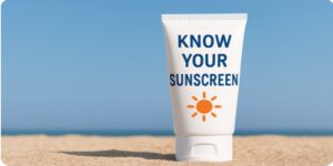 A white bottle of sunscreen standing on the sand with the words 'Know Your Sunscreen' printed on it, under a clear blue sky.